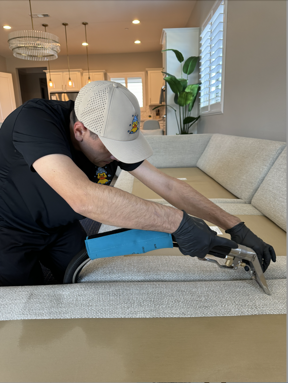 Squeaky technician in duck-logo uniform extracting dirt from a light sectional sofa